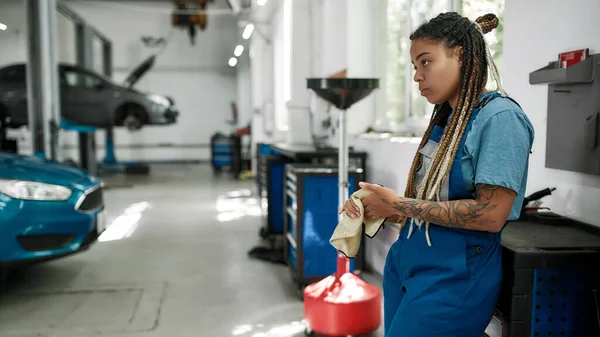Doctor para su vehículo. Mujer afroamericana joven, mecánico profesional mirando hacia otro lado, limpiando, limpiando sus manos con tela después de reparar un coche en un taller de reparación de automóviles — Foto de Stock