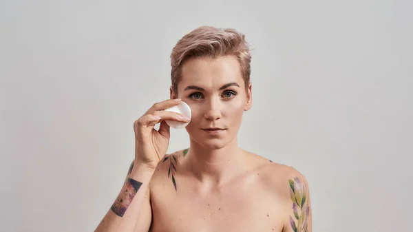 Portrait of beautiful tattooed woman with pierced nose and short hair removing makeup, cleaning skin using cotton pad isolated on grey background — Stock Photo, Image