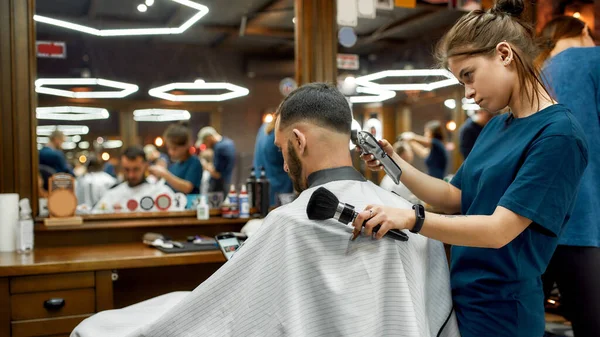 Giovane barbiera professionista che lavora con tosatrice di capelli, rendendo taglio di capelli alla moda per un giovane uomo barbuto bello in visita barbiere. Focus su donna e cliente — Foto Stock