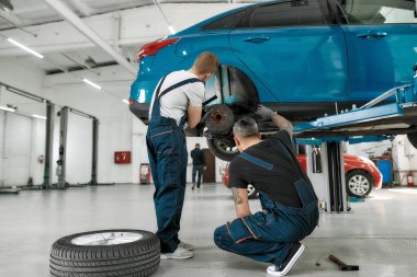 Oto bakım bizim sorumluluğumuzda. Araba tamirhanesinde kaldırılan otomobilin fren diskini incelemek için meşale kullanan iki erkek tamircinin tam boy görüntüsü.