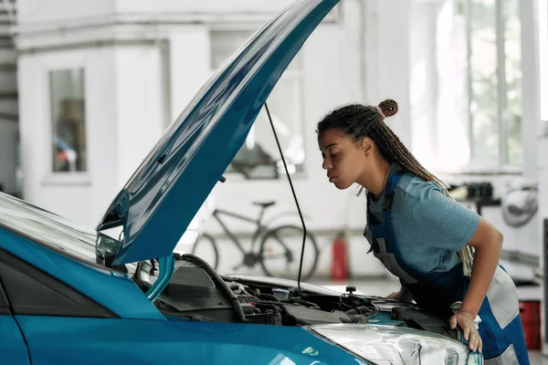 Cuidado fácil. Mujer afroamericana joven, mecánico profesional mirando, examinando debajo de la capucha del coche en el taller de reparación de automóviles — Foto de Stock