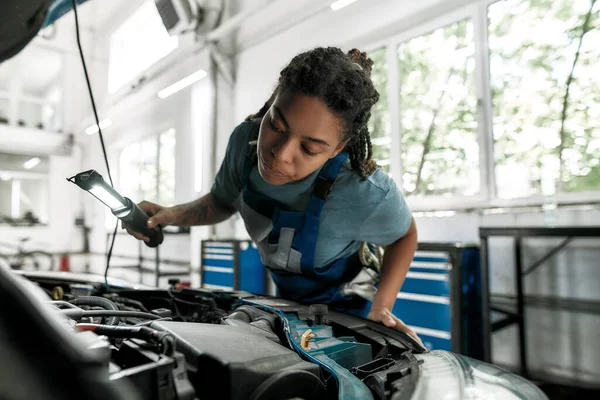 Momenten van zorg. Jonge Afro-Amerikaanse vrouw, professionele vrouwelijke monteur op zoek, onderzoek onder motorkap van auto met fakkel in auto reparatiewerkplaats — Stockfoto