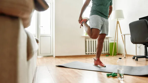 Mi sto scaldando. Colpo ritagliato di giovane uomo attivo che si allunga le gambe, facendo allenamento mattutino con manubri sul tappetino da yoga a casa — Foto Stock