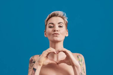 Çekici, yarı çıplak, mükemmel derili bir kadının portresi. Kameraya bakıyor. Mavi arka planda elleri izole bir kalp var.