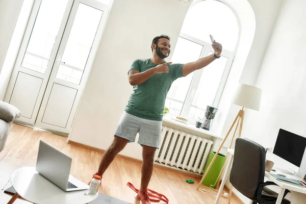 Proud. Full length shot of excited bearded active man holding smartphone, pointing while taking a selfie during morning workout at home. Fitness, motivation concept