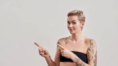 Bana bak. Kısa saçlı, yarı çıplak bir kadının portresi. Kameraya bakıyor, işaret parmaklarıyla sol tarafı işaret ediyor. Arka planda izole edilmiş.