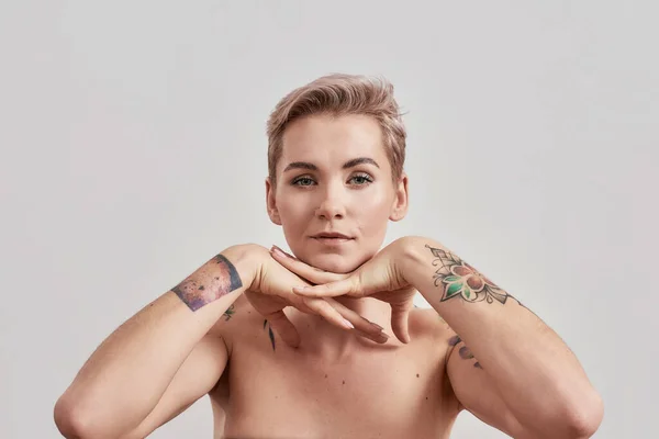 Portrait de femme tatouée à moitié nue aux cheveux courts regardant la caméra, posant les mains sous le menton isolé sur un fond clair — Photo