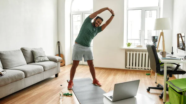Kişisel antrenör. Erkek fitness eğitmeninin evde dizüstü bilgisayar kullanarak antrenman yaparken vücudunu esnettiği uzun bir çekim. Spor, online spor salonu konsepti — Stok fotoğraf