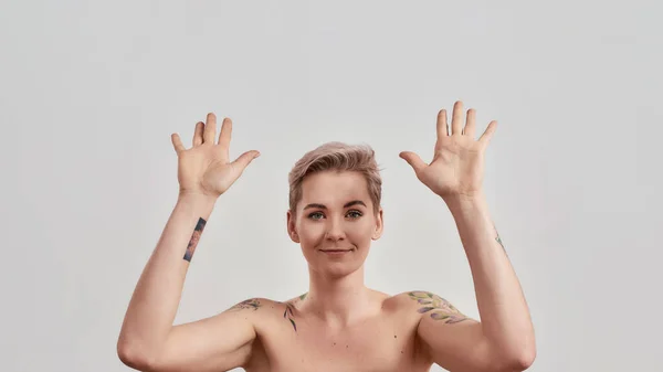 Ritratto di donna mezza nuda tatuata con i capelli corti che guarda la macchina fotografica, alzando le mani verso l'aria isolata su sfondo chiaro — Foto Stock
