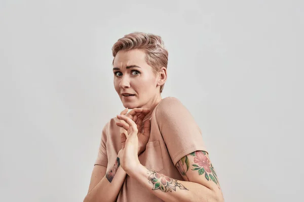 Retrato de mujer tatuada asustada con la nariz perforada y el pelo corto en camiseta beige que mira acurrucada, aterrorizada aislada sobre fondo claro — Foto de Stock