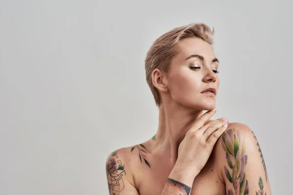 Skönhet porträtt av en ung attraktiv halvnaken tatuerad kvinna med perfekt hud tittar bort, hålla handen nära ansiktet isolerad över grå bakgrund — Stockfoto