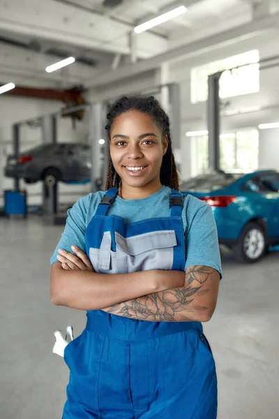 Jag är här för att tjäna dig. Porträtt av ung afrikansk amerikansk kvinna, professionell kvinnlig mekaniker i uniform ler mot kameran, står i bilverkstad. Bilservice, reparation, underhåll, personal koncept — Stockfoto