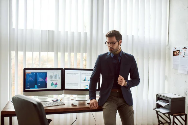 Hombre de negocios seguro en el desgaste clásico y gafas de pie cerca de su lugar de trabajo en la oficina moderna, el análisis de datos estadísticos, gráficos financieros y gráficos en la pantalla de la computadora —  Fotos de Stock