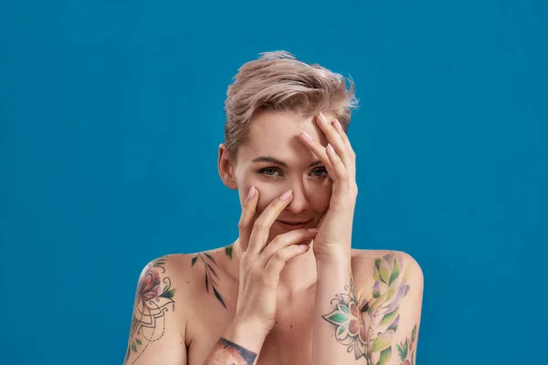 Retrato de una joven atractiva mujer semidesnuda tatuada con la piel perfecta mirando a la cámara, sosteniendo las manos sobre su cara aislada sobre fondo azul — Foto de Stock