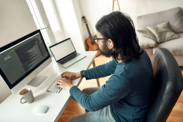 Vue latérale du jeune développeur web masculin portant des lunettes code d'écriture sur ordinateur de bureau tout en travaillant à la maison. Freelance, travail en ligne, bureau à domicile — Photo