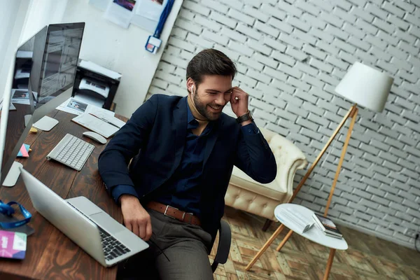 Young successful businessman or sales manager wearing wireless earphones, talking with client or partner and smiling while working in the modern office — Stock Photo, Image