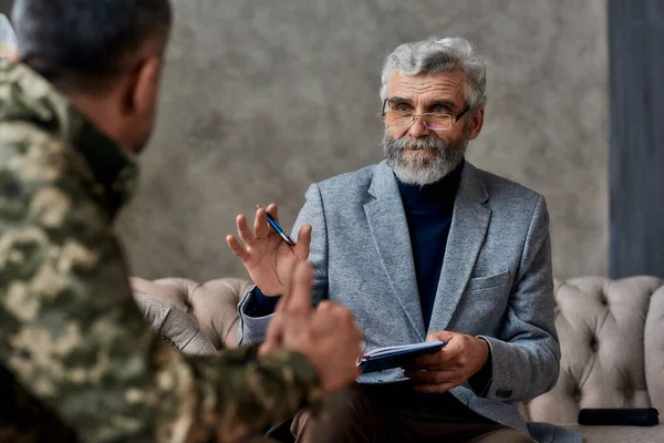 A arranjar-te. Retrato de psicólogo maduro se comunicando com o homem militar e fazendo anotações durante a sessão de terapia. Soldado que sofre de depressão, trauma psicológico. Conceito de TEPT — Fotografia de Stock