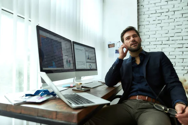 Joven hombre de negocios con auriculares inalámbricos hablando con el cliente o socio acerca de los negocios, sentado en su lugar de trabajo en la oficina moderna. Análisis de datos estadísticos —  Fotos de Stock