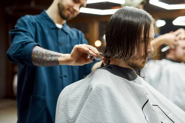 Serving client. Young hipster guy getting new trendy haircut in the modern barbershop. Professional barber cutting long hair of young man. Side view