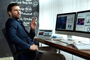 Kablosuz kulaklıklı genç iş adamı müşteri ya da ortağıyla konuşuyor, modern ofiste istatistiksel verilerle çalışırken satış raporlarını tartışıyor.