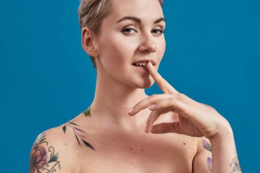 Genç, çekici, yarı çıplak, derisi dövmeli, neşeli görünen, parmaklarını ağzında tutan ve mavi arka planda izole bir şekilde gülümseyen bir kadının portresini yakından çek.