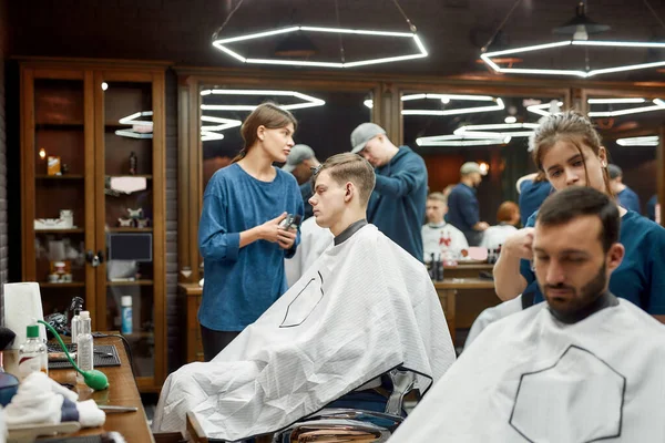 Gente en la barbería. Joven peluquera profesional, peluquera femenina que trabaja con el cortador de pelo, sirviendo chico guapo joven sentado en la silla de la barbería — Foto de Stock