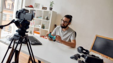 Gözlüklü bir erkek teknoloji blogcusu oyun kumandası joystick 'e bakıyor, video blogu veya evdeki yeni cihazlar hakkında video blogu kaydederken onu tutuyor