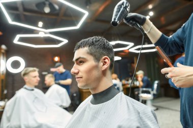 Çalışma süreci. Modern berber dükkanında yakışıklı bir adamın saçını kurutan berberin yan görüntüsü.