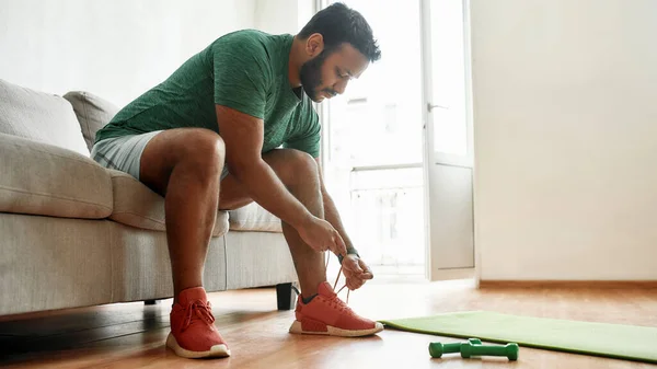 Casi listo. Joven hombre activo en zapatillas de deporte atando cordones, preparándose para el entrenamiento matutino con pesas en una esterilla de yoga en casa. Fitness, concepto de motivación — Foto de Stock