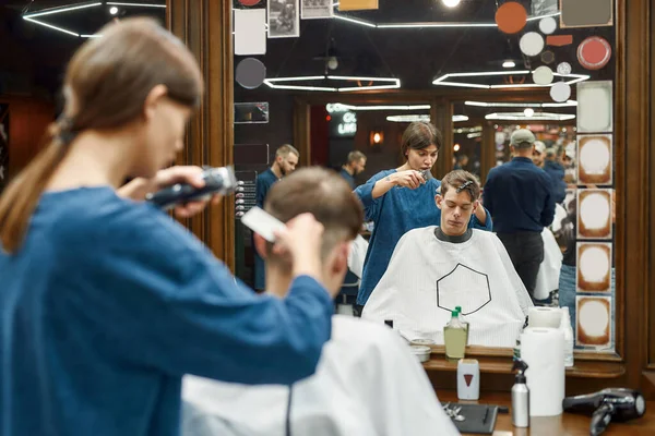 Młoda, profesjonalna fryzjerka pracująca z maszynką do strzyżenia, obsługująca młodego faceta siedzącego w fotelu fryzjerskim przed lustrem. Skupienie selektywne — Zdjęcie stockowe