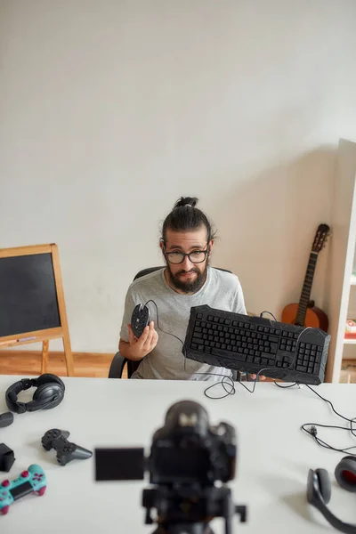 Blogger de tecnología masculina en gafas sosteniendo, mostrando el teclado y el ratón mientras graba video blog o vlog sobre nuevos dispositivos en casa —  Fotos de Stock