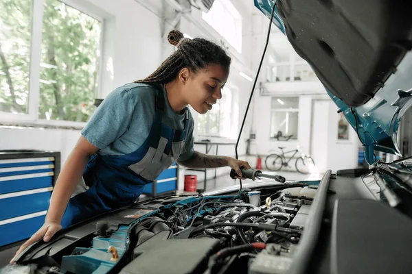 Oprava se povedla. Mladý africký Američan žena, profesionální žena mechanik hledá, zkoumá pod kapotou auta s baterkou v autoopravně — Stock fotografie