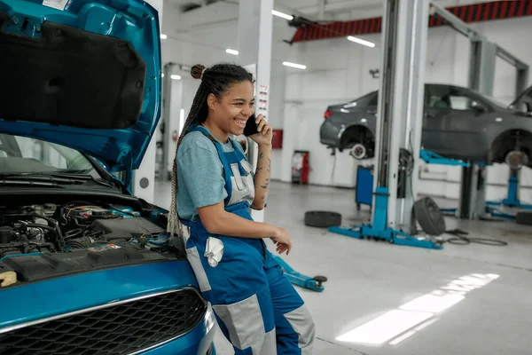 Young african american woman, professional female mechanic talking on phone, leaning on a car with open hood at auto repair shop — Stock Photo, Image