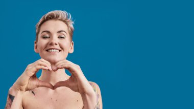 Mükemmel ciltli, yarı çıplak, genç bir kadının portresi. Mutlu görünüyor. Mavi arka planda elleri izole bir kalp var.