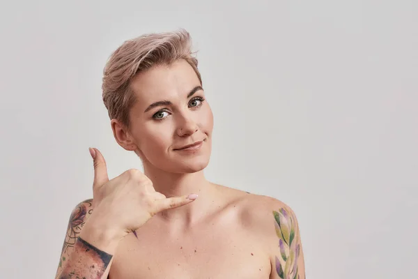 Chiamami. Ritratto di donna mezza nuda tatuata con i capelli corti gesticolare una telefonata con mano, guardando la fotocamera isolata su sfondo chiaro — Foto Stock