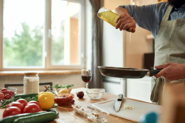 Vágott felvétel az emberről, kötényben főz, olajat önt a serpenyőre, hogy elkészítse a vacsorát, a konyhában áll. Főzés otthon koncepció — Stock Fotó