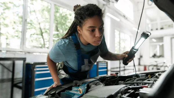 Trochu péče o vaše vozidlo. Mladý africký Američan žena, profesionální žena mechanik hledá, zkoumá pod kapotou auta s baterkou v autoopravně — Stock fotografie