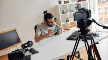 Gözlüklü bir erkek teknoloji blogcusu oyun kumandası joystick 'e bakıyor, video blogu veya evdeki yeni cihazlar hakkında video blogu kaydederken onu tutuyor