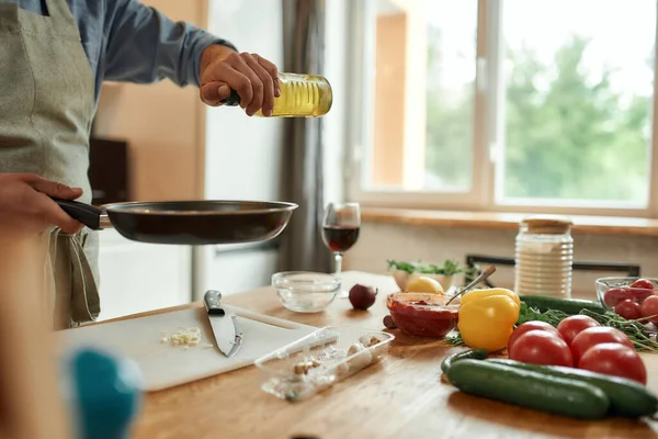 Vágott felvétel az emberről, kötényben főz, olajat önt a serpenyőre, hogy elkészítse a vacsorát, a konyhában áll. Főzés otthon koncepció — Stock Fotó