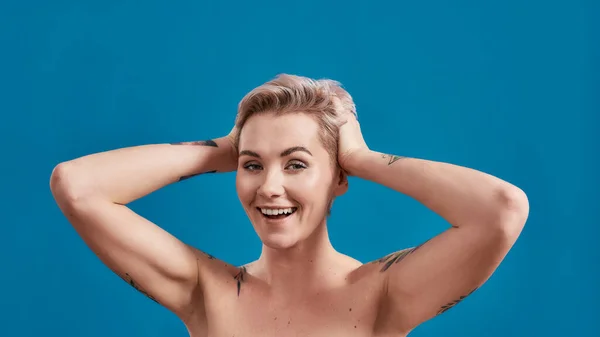 Beauty portrait of a young attractive half naked tattooed woman with perfect skin looking excited, arms folded behind her head isolated over blue background