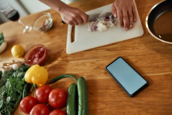 Κλείσιμο τηλεφώνου με κενή οθόνη στο τραπέζι. Άνθρωπος κοπής κρεμμυδιού σε ξύλο κοπής, χρησιμοποιώντας την εφαρμογή smartphone, ενώ το μαγείρεμα δείπνο. Υγιεινή διατροφή, τεχνολογική έννοια — Φωτογραφία Αρχείου