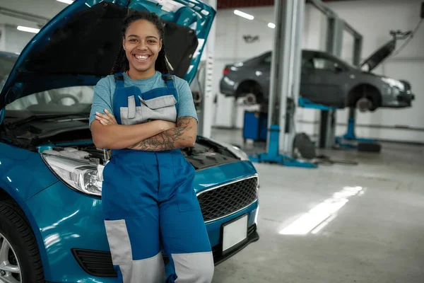 Mantén tu auto en movimiento. Mujer afroamericana joven, mecánico profesional sonriendo a la cámara, apoyándose en un coche en el taller de reparación de automóviles — Foto de Stock