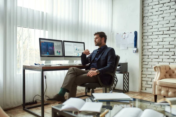 Full length of young thoughtful businessman or financial analyst sitting at his workplace in the modern office, looking at graphs and charts on pc screen and thinking about business — Stock Photo, Image