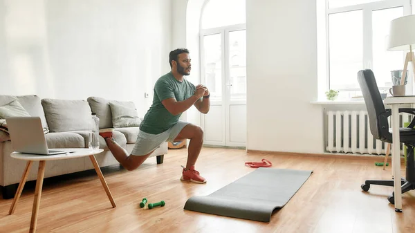 Get Fit At Home. Full length shot of young active man watching online video training on laptop, exercising, stretching during morning workout at home. Sport, healthy lifestyle