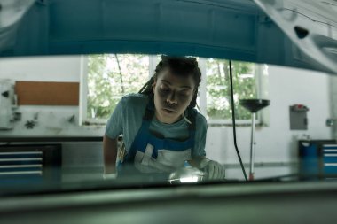 Genç Afro-Amerikan kadın, profesyonel kadın tamirci görünümlü, araba tamirhanesinde el feneriyle arabanın kaputunu inceliyor.