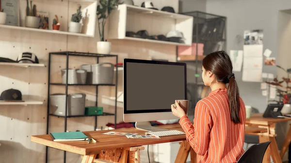 Aventuras na criatividade. Retrato de trabalhadora sentada ao lado da mesa, bebendo café enquanto desenha esboços design do logotipo usando pc. Jovem que trabalha na T-shirt personalizada, empresa de impressão de roupas — Fotografia de Stock