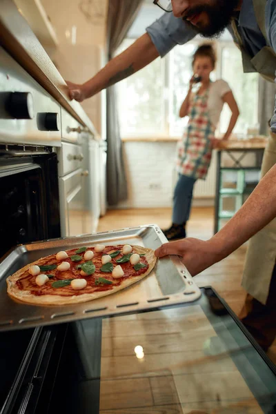 Cocinero profesional haciendo pizza en casa. Hombre poniendo pizza cruda en el horno moderno para hornear. Mujer de pie en el fondo. Hobby, estilo de vida — Foto de Stock