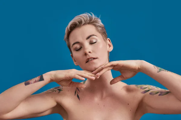 Retrato de una joven atractiva mujer semidesnuda tatuada con la piel perfecta cerrando los ojos, posando con las manos cerca de su cara, tocando la barbilla aislada sobre fondo azul — Foto de Stock