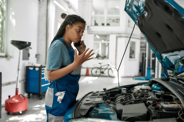 Servicio personalizado. Mujer afroamericana joven, mecánico profesional hablando por teléfono emocionalmente, mientras examina el motor bajo la capucha del coche en el taller de reparación de automóviles — Foto de Stock