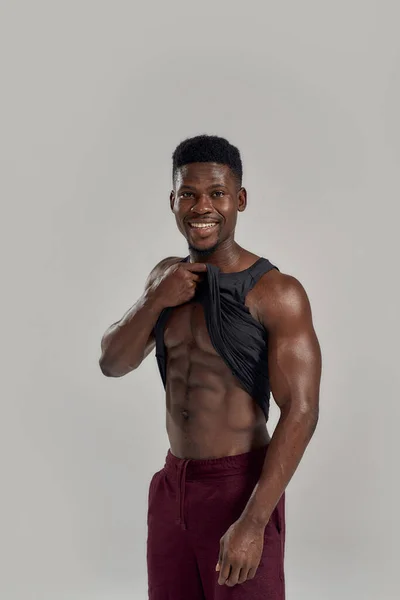 Jovem musculoso afro-americano sorrindo para a câmera, levantando camisa preta, mostrando seus abdominais enquanto posava isolado sobre fundo cinza. Esportes, treino, conceito de musculação — Fotografia de Stock